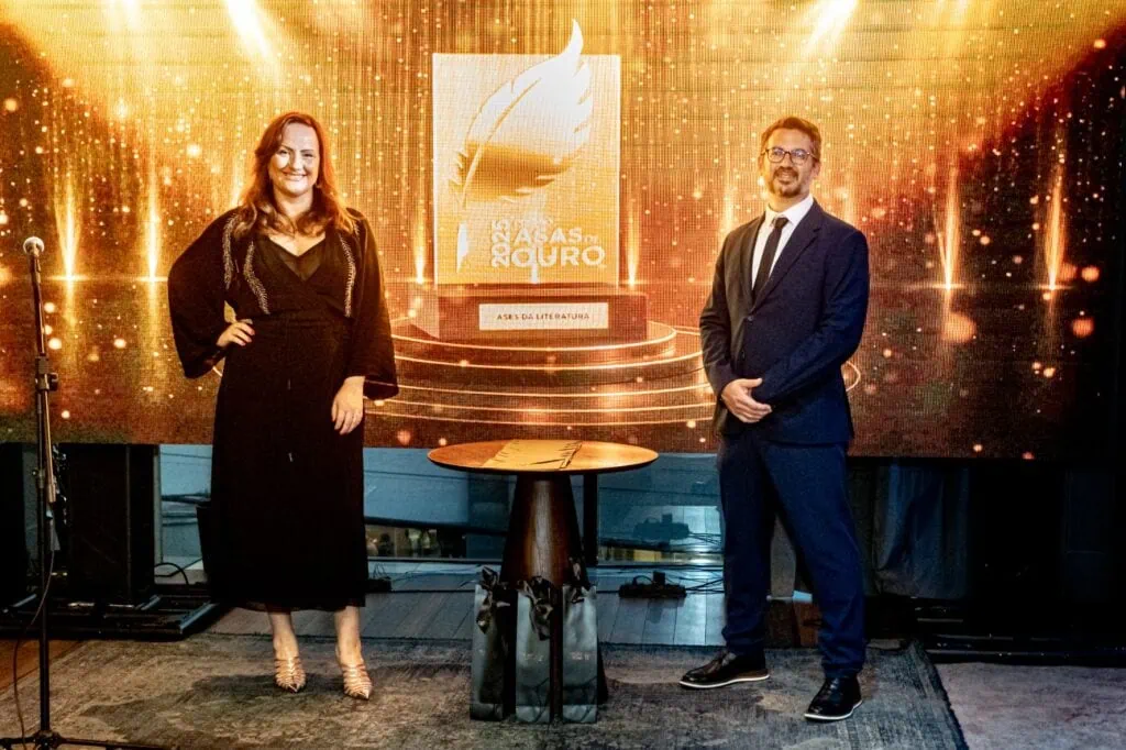 Dois participantes ao lado da mesa de troféus, diante do painel dourado do Prêmio Asas de Ouro